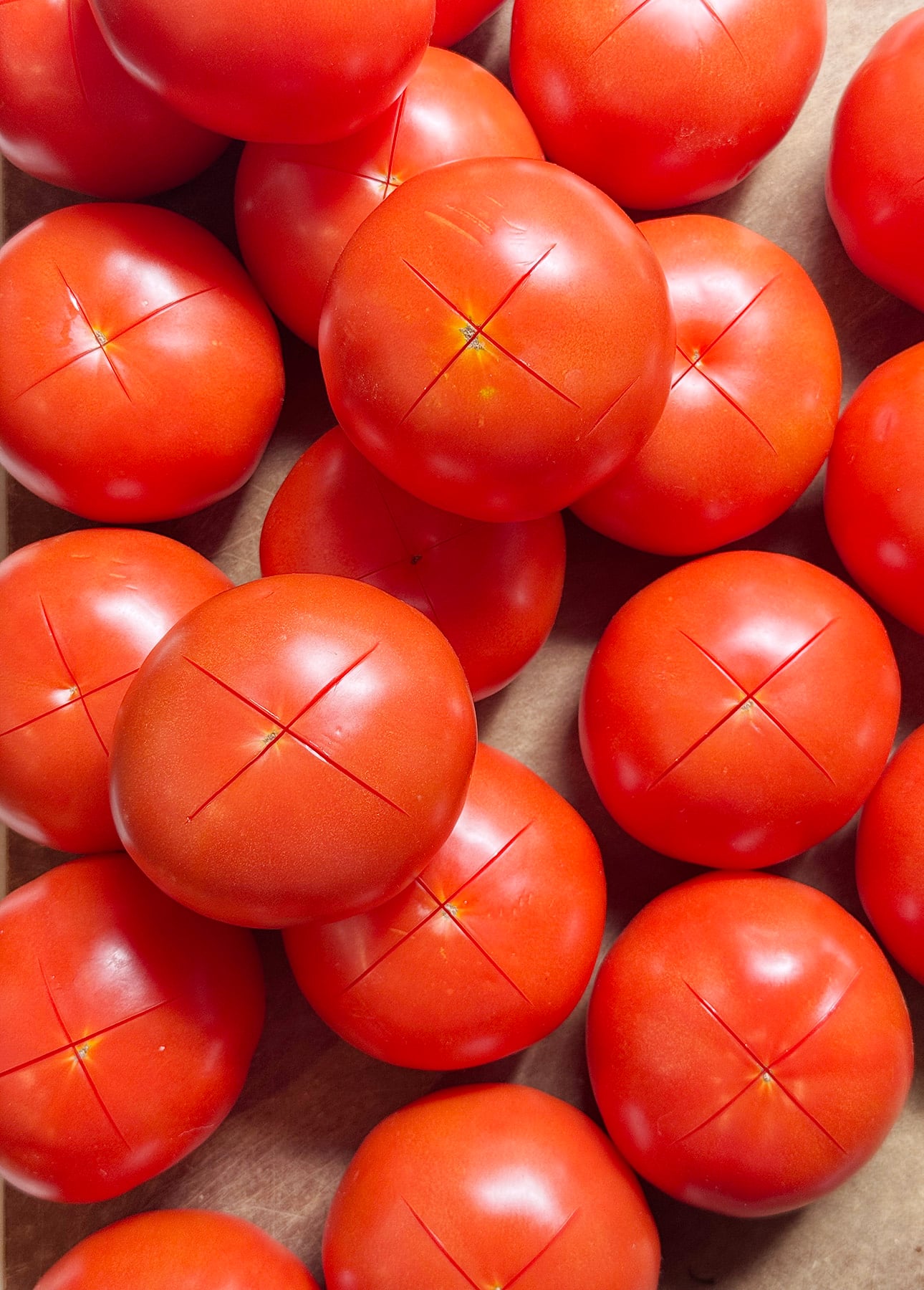 A group of red tomatoes with shallow X-shaped cuts on their tops, arranged closely together on a brown surface—ideal for making a vibrant Fresh Tomato Sauce. // FoodNouveau.com