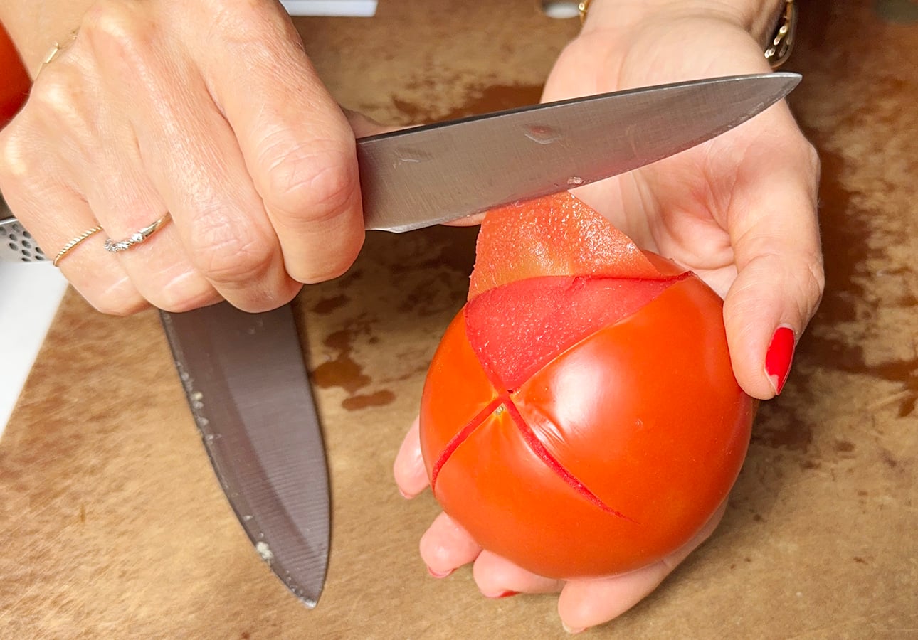 A person uses a knife to score an X on the bottom of a red tomato, preparing it for peeling—a classic step in making fresh tomato sauce. The tomato is held in one hand on a brown cutting board. // FoodNouveau.com