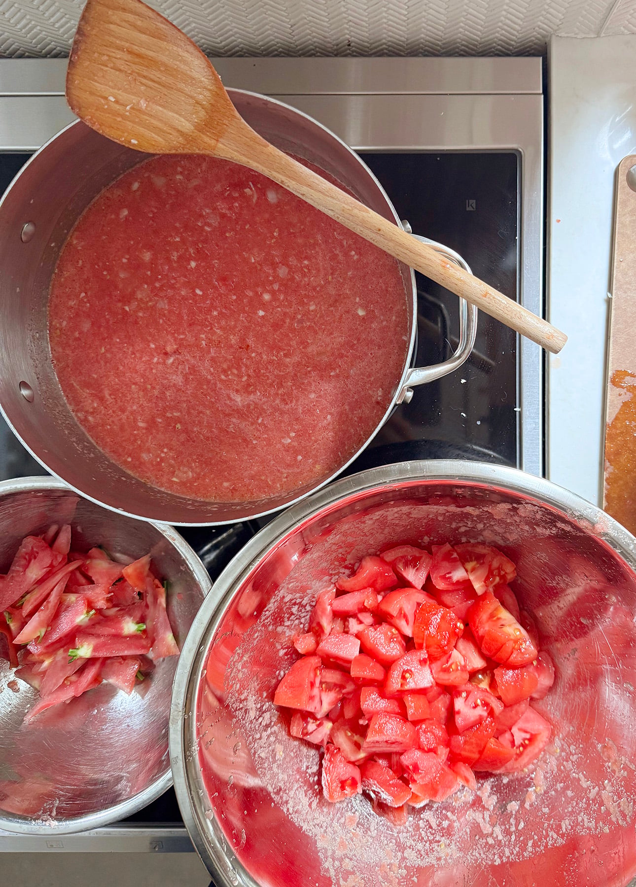Diced tomatoes in a metal bowl, tomato skins in another, and a pot of blended tomatoes simmering on the stove with a wooden spoon resting on top—all items are being prepared for cooking a fresh tomato sauce. // FoodNouveau.com