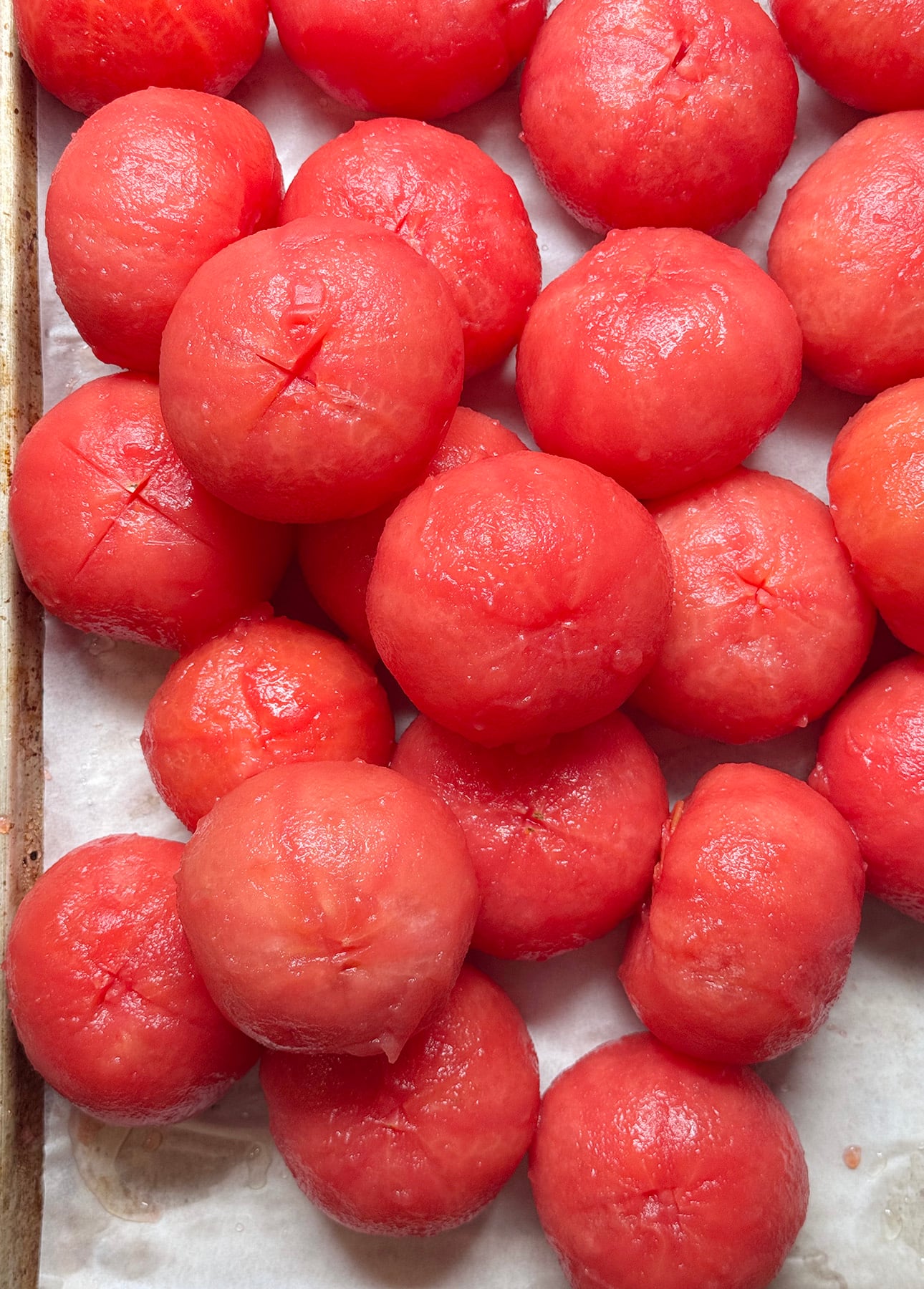 Several peeled, bright red tomatoes—perfect for fresh tomato sauce—are arranged closely together on a parchment-lined baking sheet. Their smooth surfaces and juicy texture are clearly visible. // FoodNouveau.com