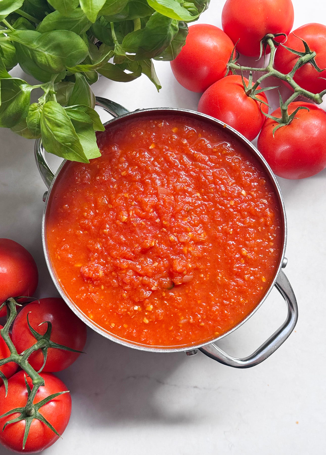 A pot of chunky Fresh Tomato Sauce sits on a white surface, surrounded by fresh basil leaves and vine-ripened tomatoes. // FoodNouveau.com