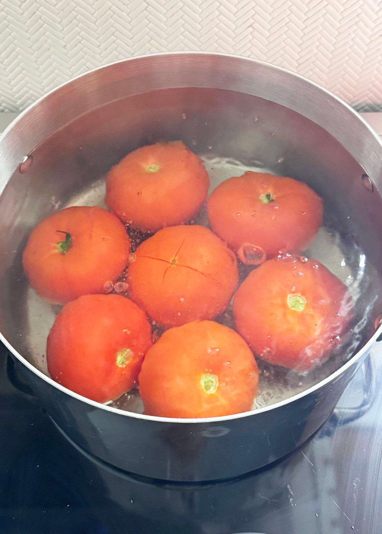 Seven tomatoes are boiling in a pot of hot water on a stove, preparing the base for a delicious Fresh Tomato Sauce. One tomato has an X cut into its skin, likely to make peeling easier. The background shows a white textured wall. // FoodNouveau.com