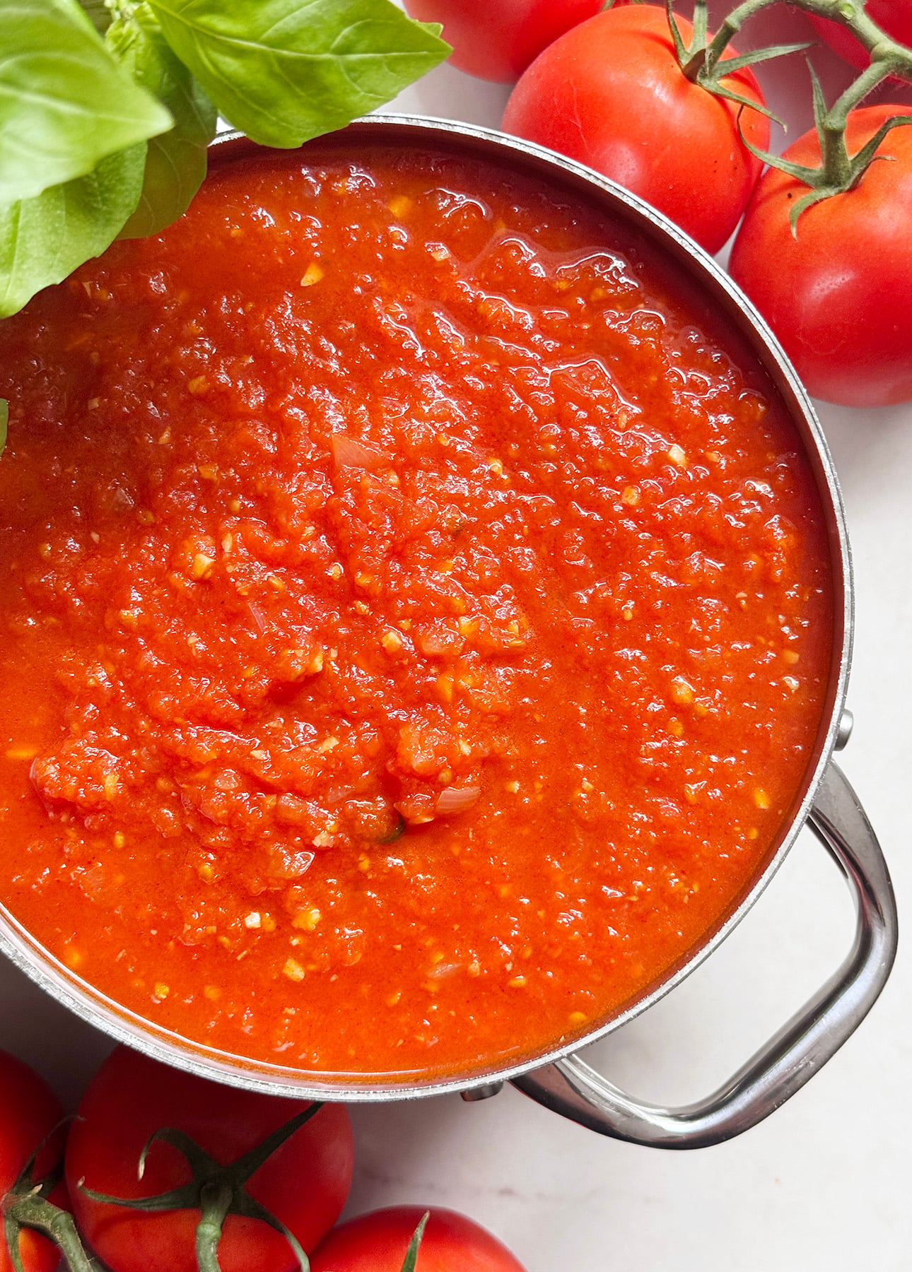 A pot of chunky fresh tomato sauce sits on a white surface, surrounded by fresh tomatoes on the vine and basil leaves. // FoodNouveau.com