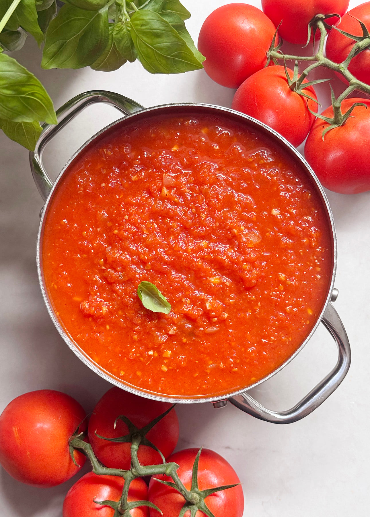 A pot of fresh tomato sauce garnished with a basil leaf, surrounded by fresh tomatoes on the vine and basil leaves on a white surface. // FoodNouveau.com