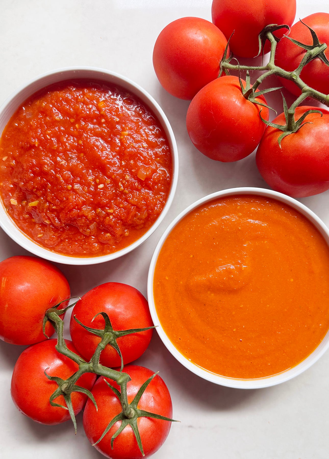 Two bowls of fresh tomato sauce, one chunky and one smooth, are surrounded by vibrant vine tomatoes on a white surface. // FoodNouveau.com