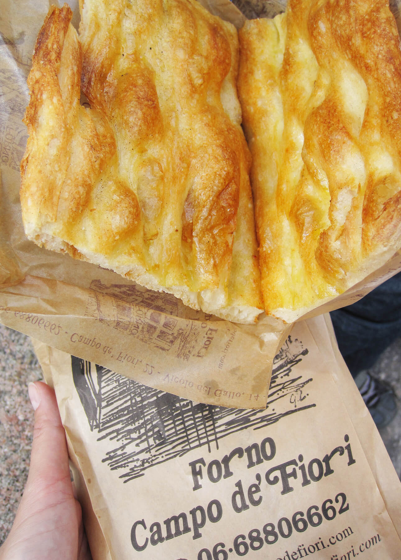 A hand holds a piece of golden, airy focaccia bread&mdash;reminiscent of Roman-Style Pizza&mdash;on brown paper packaging labeled Forno Campo de&rsquo; Fiori, complete with contact details and a simple bakery sketch. // FoodNouveau.com