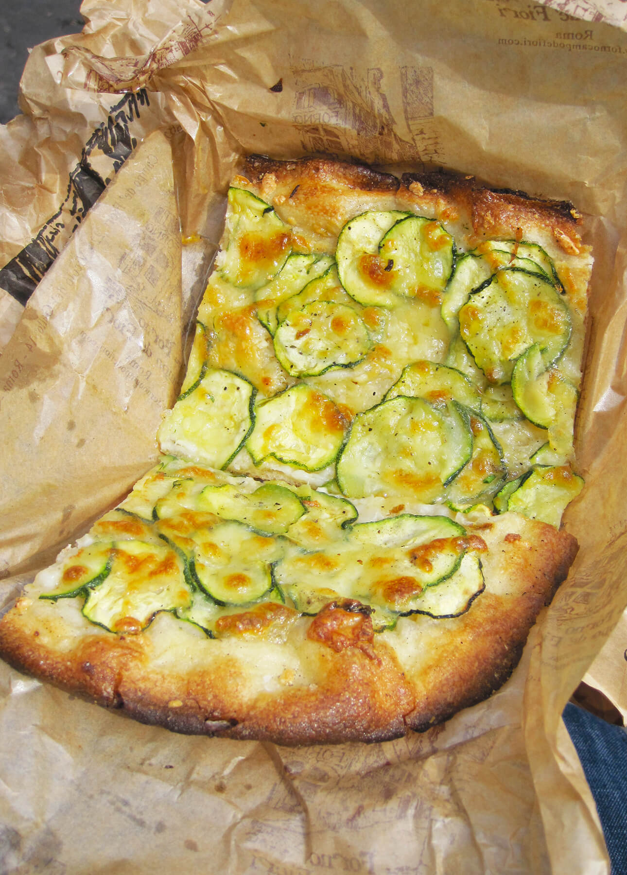 A rectangular slice of Roman-Style Pizza with melted cheese and thinly sliced zucchini on top, served on brown parchment paper. The crust is golden and slightly crisp. // FoodNouveau.com