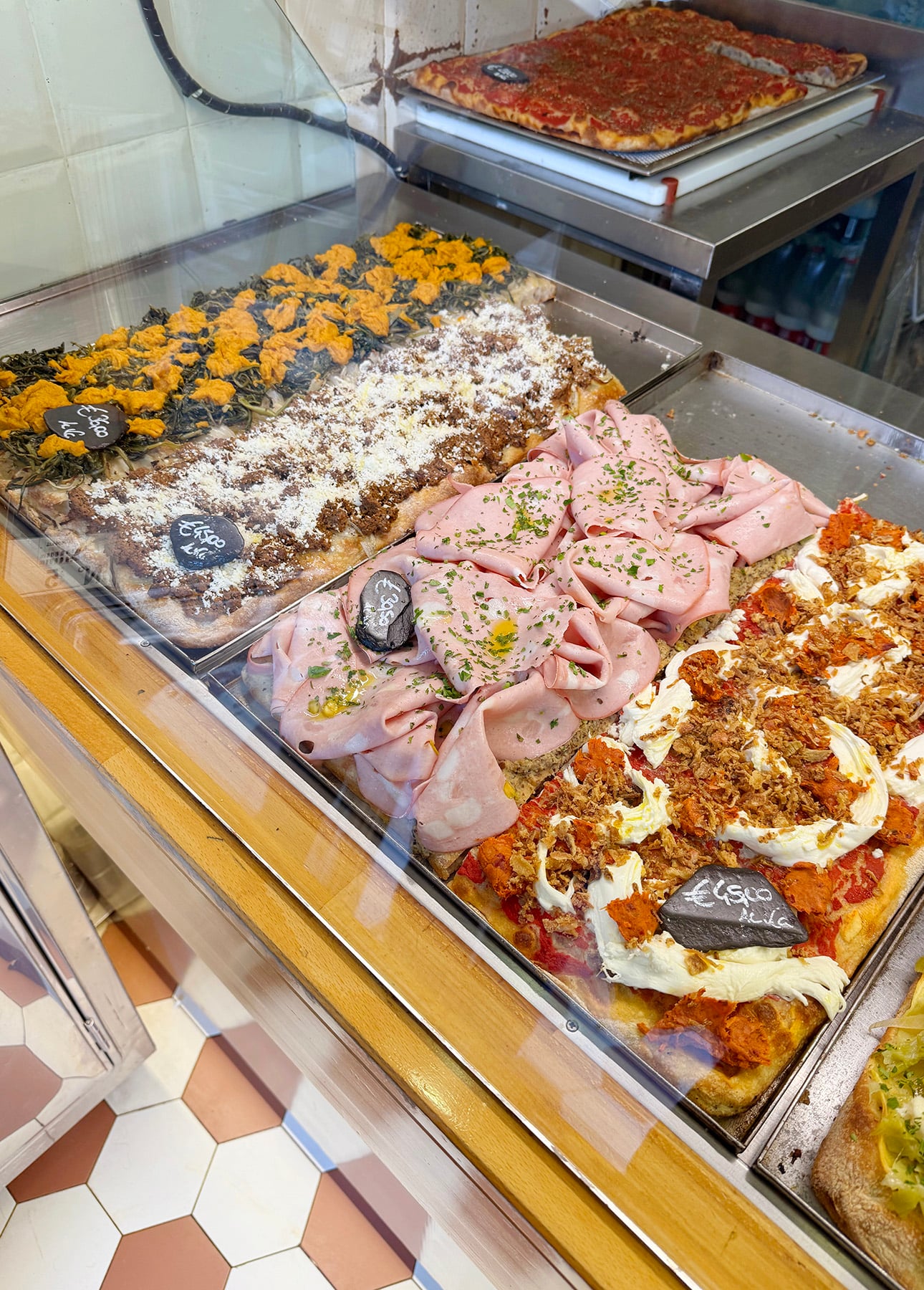 Various rectangular Roman-Style pizzas with different toppings&mdash;meats, greens, and cheese&mdash;are displayed behind a glass counter in a pizzeria. Handwritten signs label each pizza. The floor has a tile pattern. // FoodNouveau.com