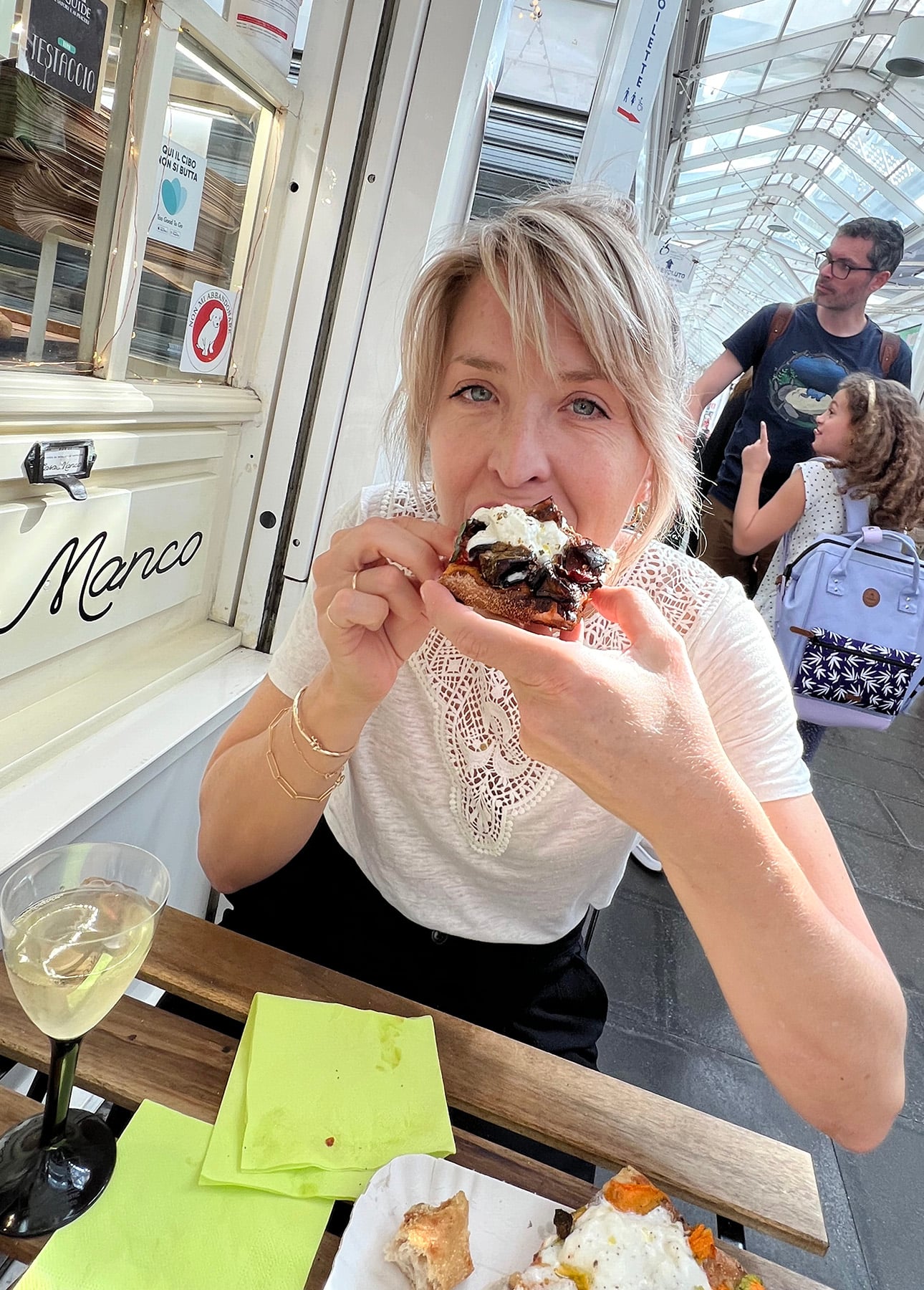A woman sits at an outdoor caf&eacute; table, smiling as she enjoys a pastry topped with cream. On the table are a glass of white wine, Roman-Style Pizza, and other food, while people stroll by in the background. // FoodNouveau.com