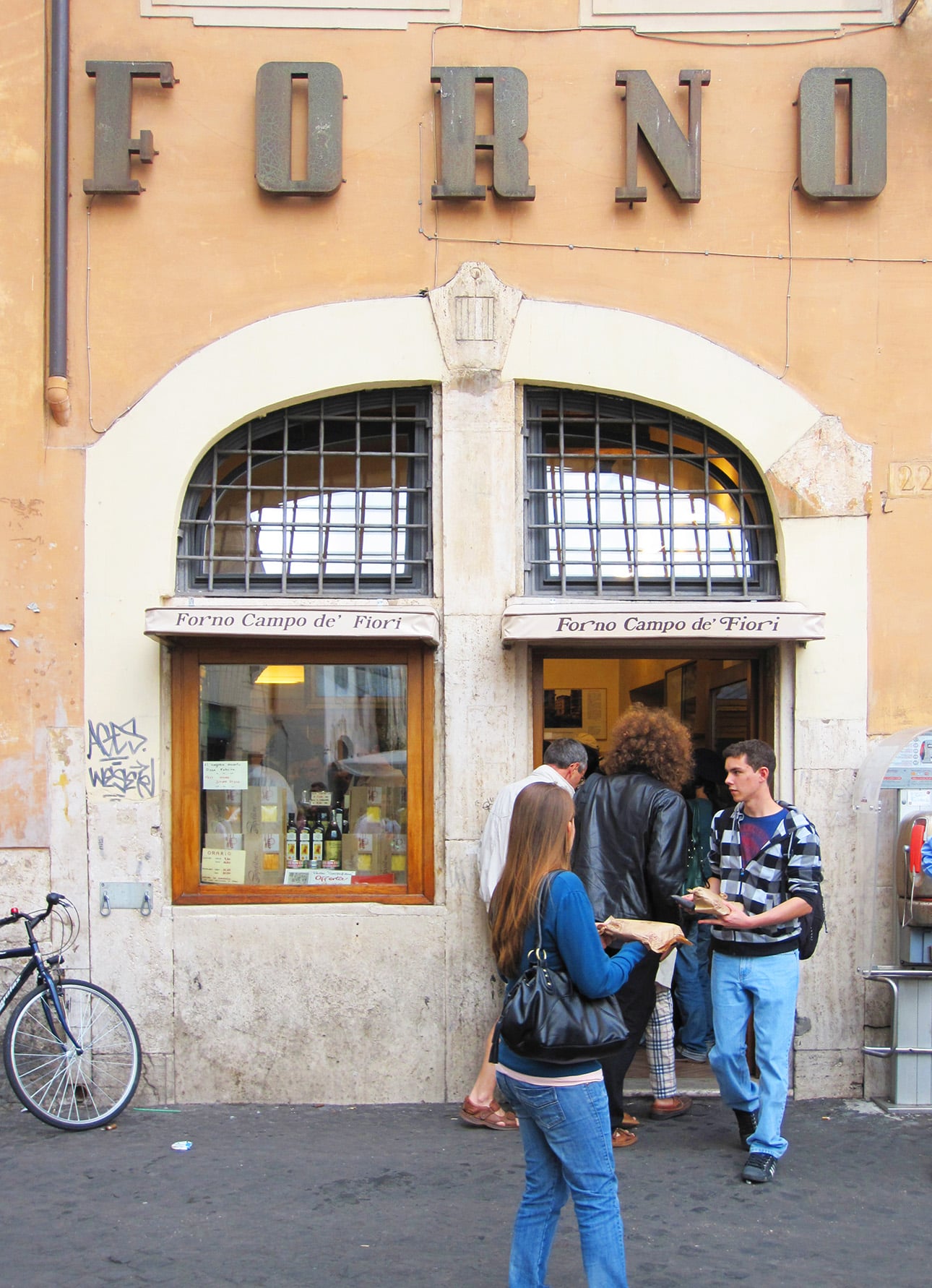 People stand outside a bakery called Forno Campo de&rsquo; Fiori, with two display windows and an arched doorway. A bicycle is parked nearby, and one woman enjoys Roman-style pizza while others queue at the entrance. // FoodNouveau.com