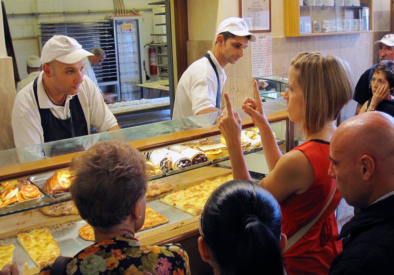 A woman in a red dress points with both index fingers at Roman-Style Pizza and pastries behind a glass counter in a busy bakery, while two staff members in white hats assist customers. Several people wait in line. // FoodNouveau.com