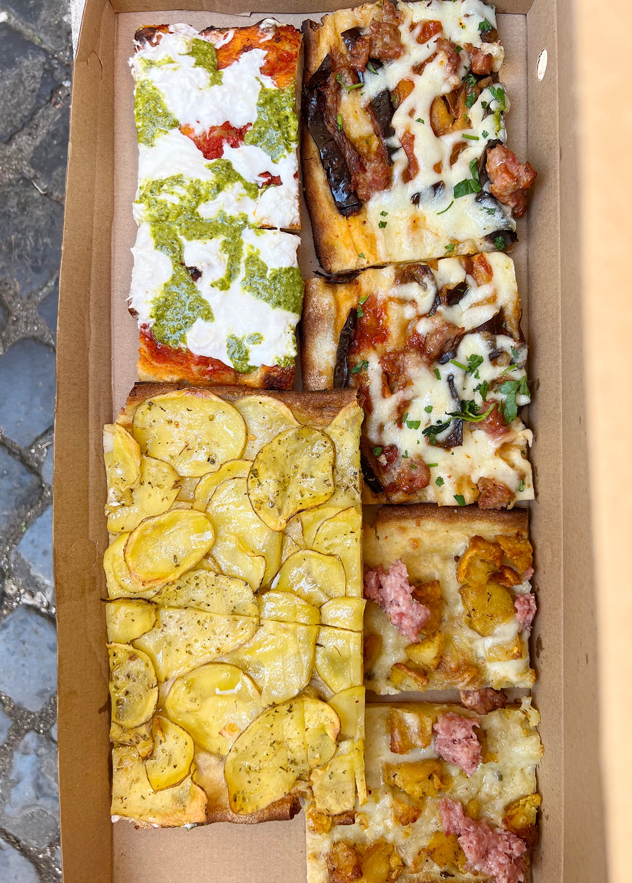 A cardboard box contains four slices of Roman-Style Pizza: one with ricotta, pesto, and tomato sauce; one with cheese, mushrooms, and herbs; one topped with thin potato slices; and another with sausage and roasted potatoes. // FoodNouveau.com