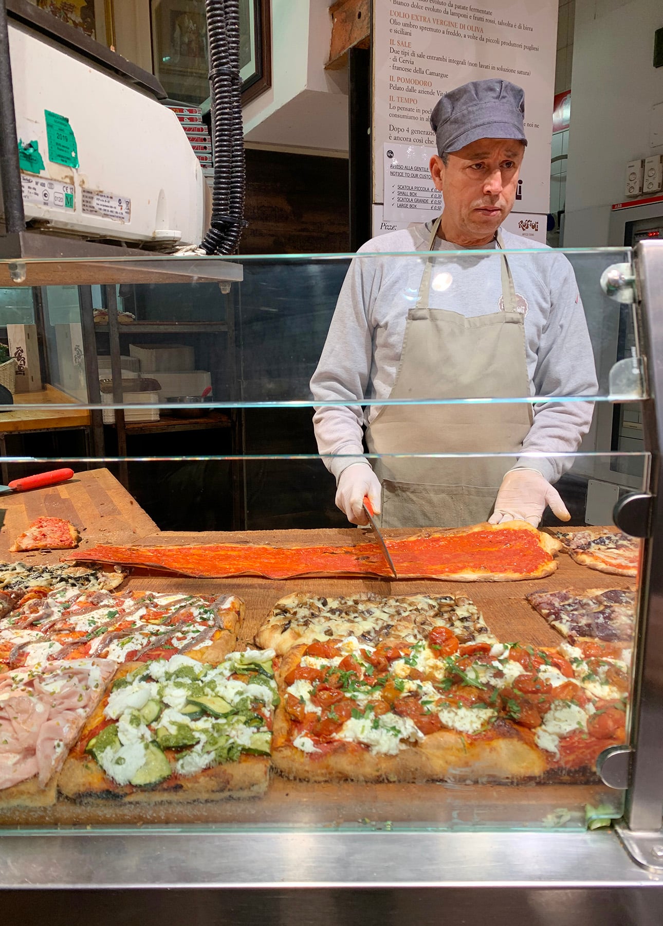 A man wearing a gray hat and apron slices a long rectangular Roman-Style Pizza with tomato sauce behind a glass counter displaying various types of square pizzas with different toppings. // FoodNouveau.com