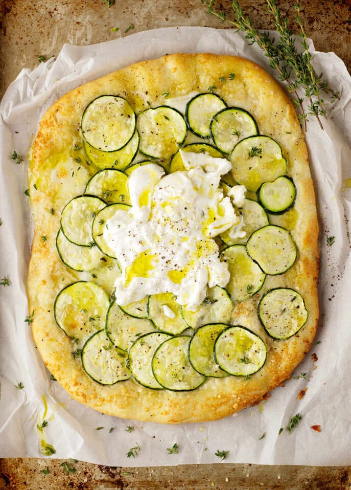 A Roman-Style Pizza featuring a rectangular flatbread topped with thinly sliced zucchini, fresh burrata cheese in the center, a drizzle of olive oil, cracked black pepper, and fresh thyme, all served on parchment paper. // FoodNouveau.com