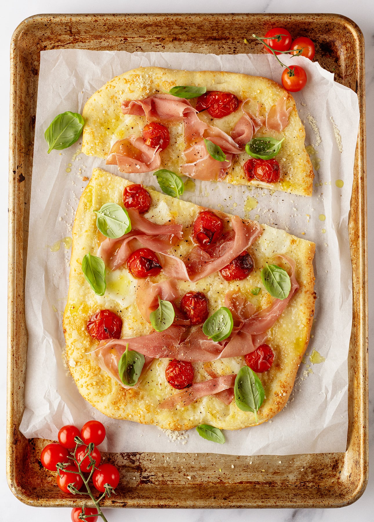 A rectangular baking tray with parchment paper holds a Roman-Style Pizza topped with cherry tomatoes, prosciutto slices, melted cheese, and fresh basil leaves. A small bunch of cherry tomatoes rests in the tray's corner. // FoodNouveau.com