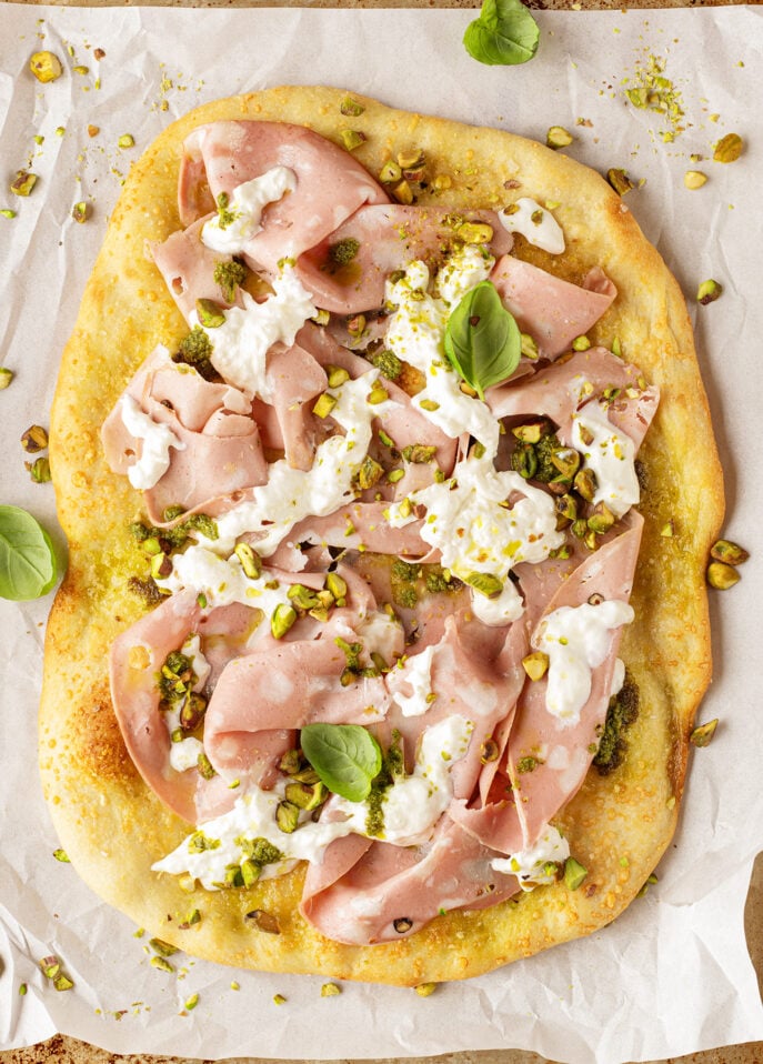 A rustic Roman-Style Pizza flatbread topped with layers of thinly sliced mortadella, creamy cheese dollops, chopped pistachios, pesto, and fresh basil leaves on parchment paper. // FoodNouveau.com