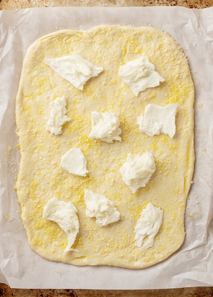 A rectangular sheet of uncooked dough on parchment paper is sprinkled with grated cheese and dotted with several dollops of ricotta cheese, ready to become a delicious Roman-Style Pizza in the oven. // FoodNouveau.com