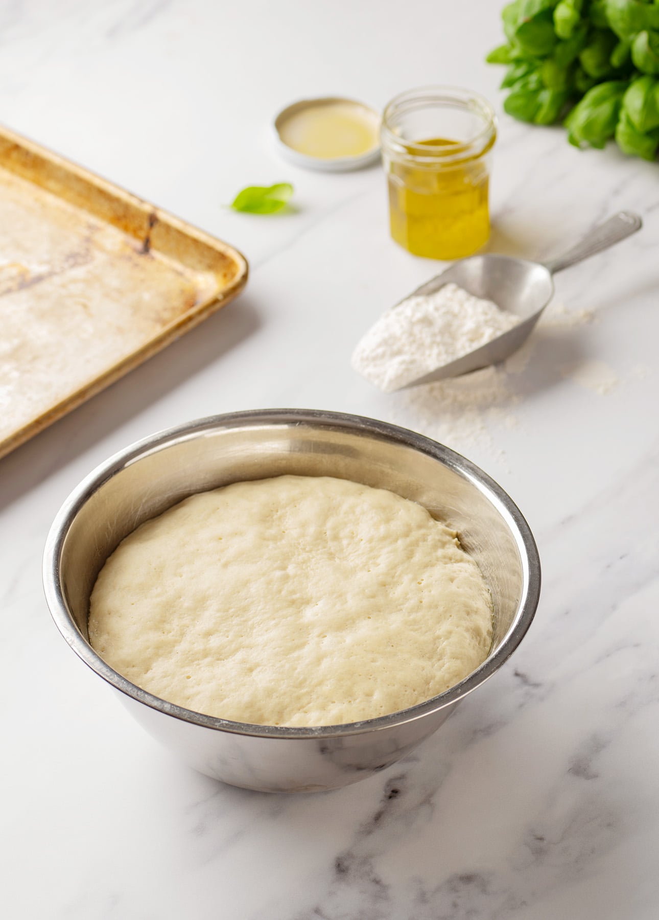 A metal bowl of risen Roman-Style Pizza dough sits on a marble countertop near a flour scoop, a jar of olive oil, a baking sheet, and fresh basil leaves. // FoodNouveau.com