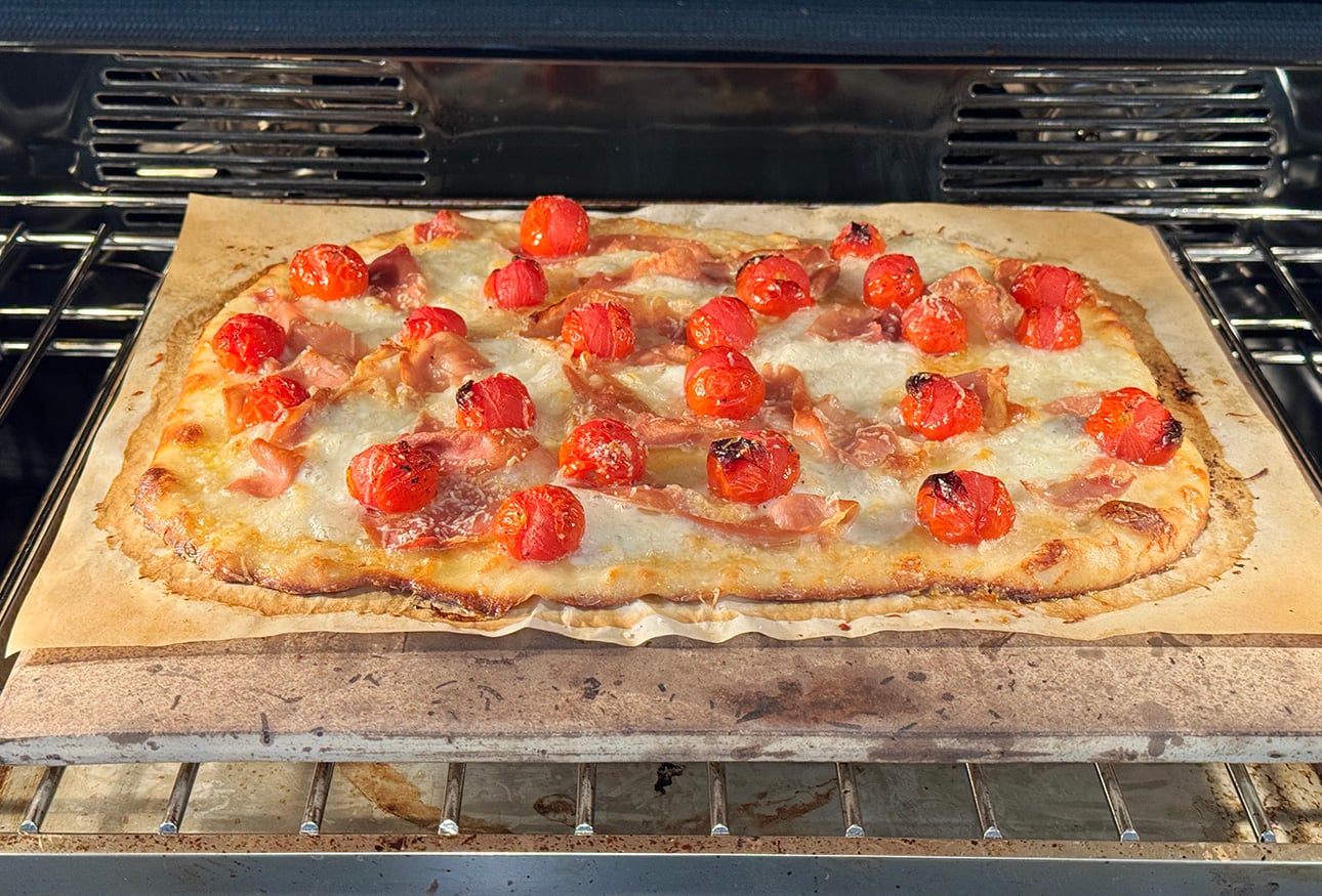 Rectangular Roman-Style Pizza topped with melted cheese, halved cherry tomatoes, and thin pieces of prosciutto, baking on parchment paper in an oven. // FoodNouveau.com