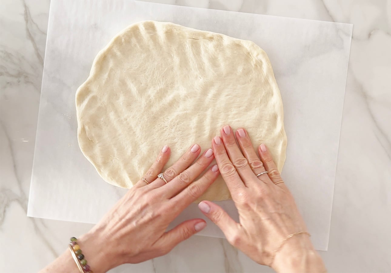 Two hands are pressing and shaping Roman-Style Pizza dough on a sheet of parchment paper placed on a marble surface. The person is wearing rings and a bracelet. // FoodNouveau.com