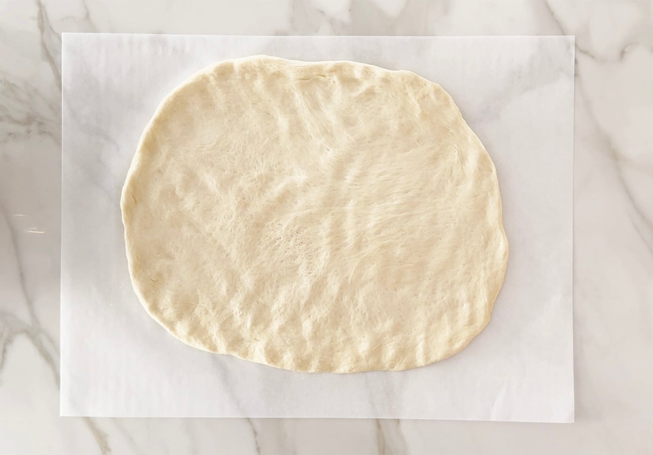 A large, oval-shaped piece of raw Roman-Style Pizza dough is spread out on a sheet of parchment paper, placed on a white marble surface. // FoodNouveau.com