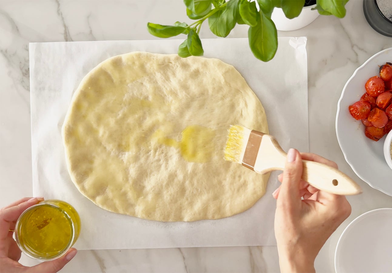 A person brushes olive oil onto rolled-out Roman-Style Pizza dough on parchment paper. Nearby are a bowl of cherry tomatoes, a green basil plant, and a small glass jar of oil on a marble countertop. // FoodNouveau.com