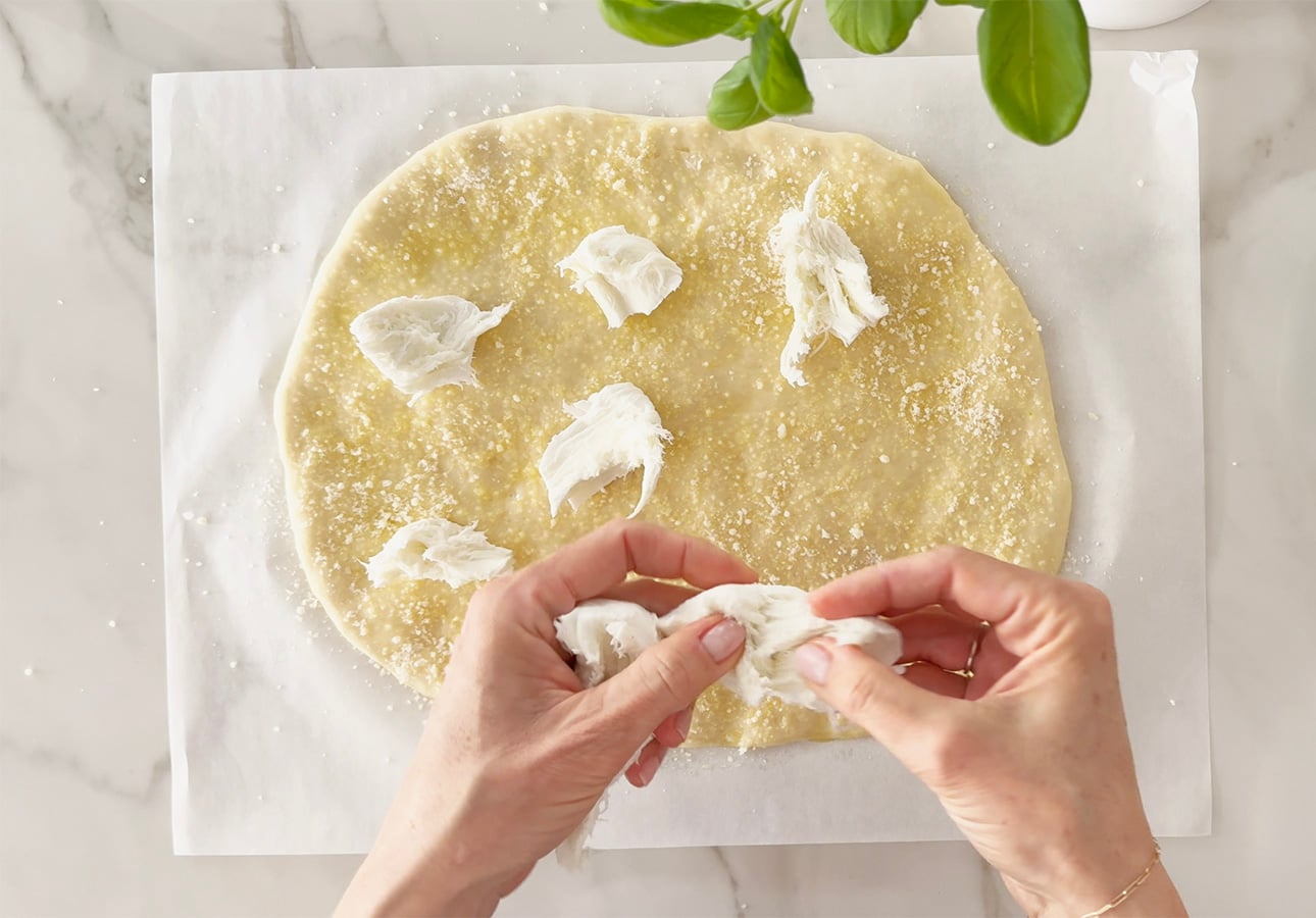 Hands tearing mozzarella cheese and placing pieces on a round, uncooked Roman-Style Pizza dough sprinkled with flour, on parchment paper over a marble surface. Green basil leaves are visible at the top of the image. // FoodNouveau.com