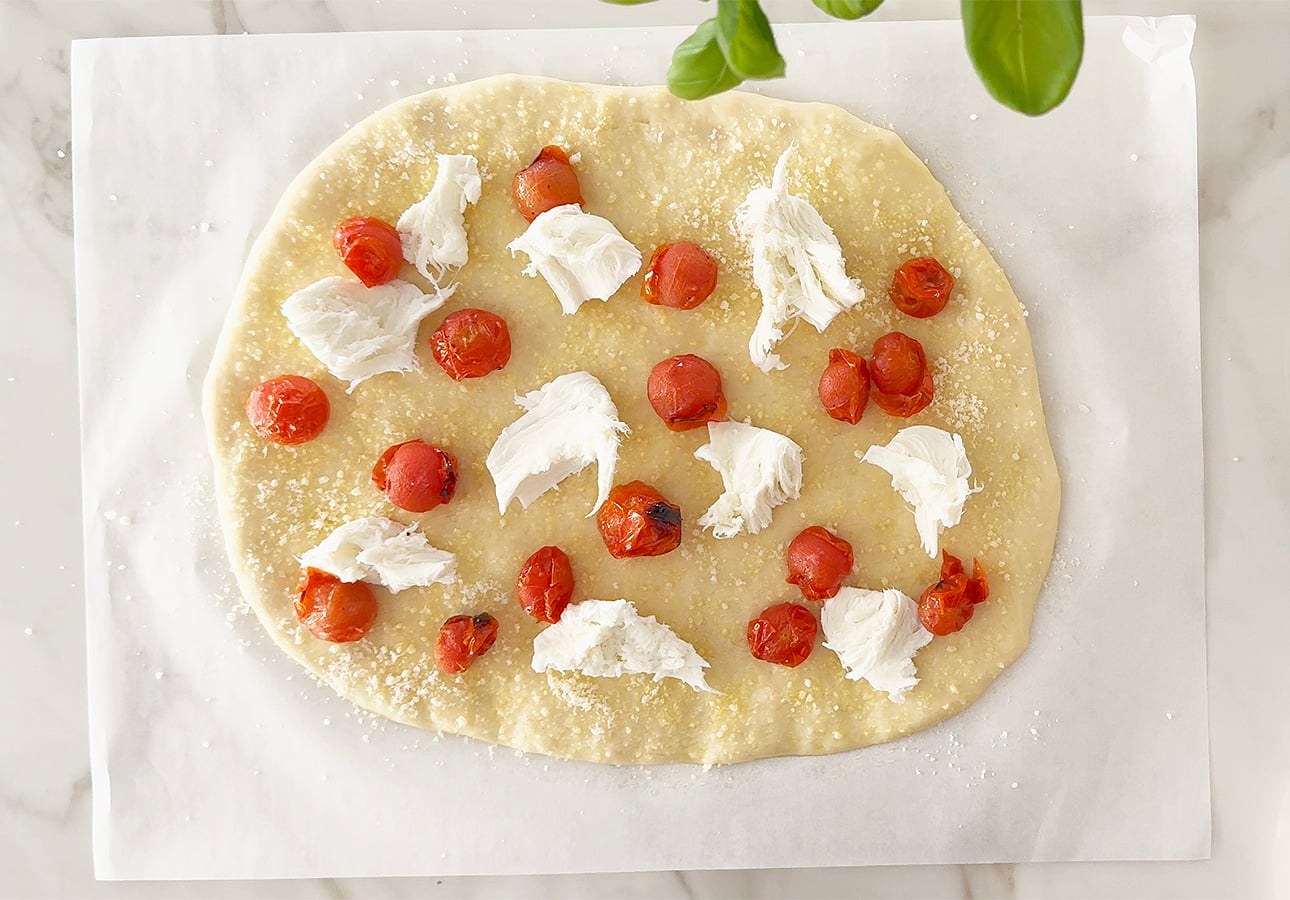 An uncooked Roman-Style Pizza base topped with cherry tomatoes, mozzarella cheese pieces, and grated Parmesan sits on parchment paper on a marble surface. Green basil leaves hang above the pizza. // FoodNouveau.com