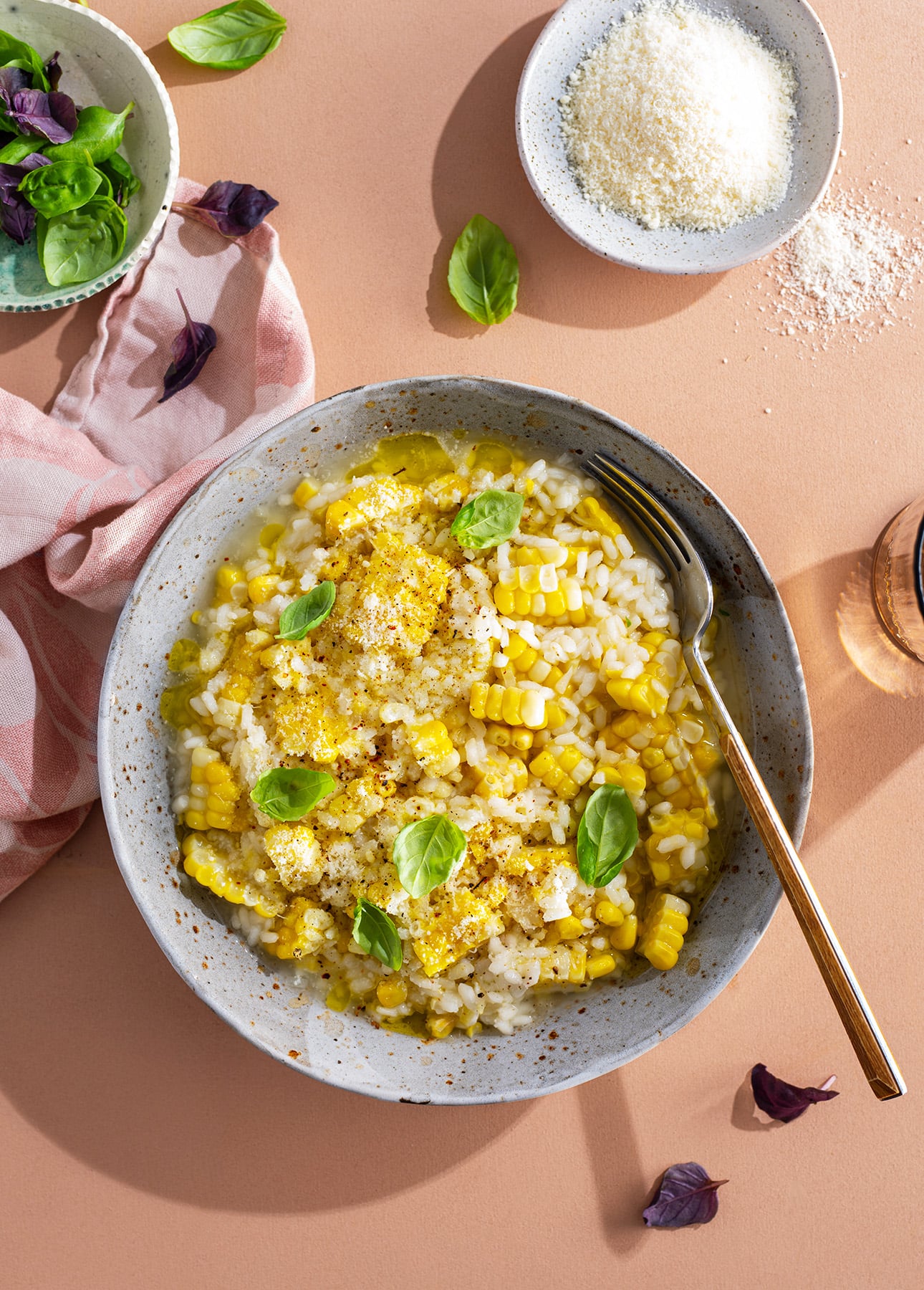 A bowl of creamy corn risotto topped with grated cheese and fresh basil leaves sits on a peach-colored surface beside a fork, a bowl of grated cheese, and a small bowl of greens. // FoodNouveau.com