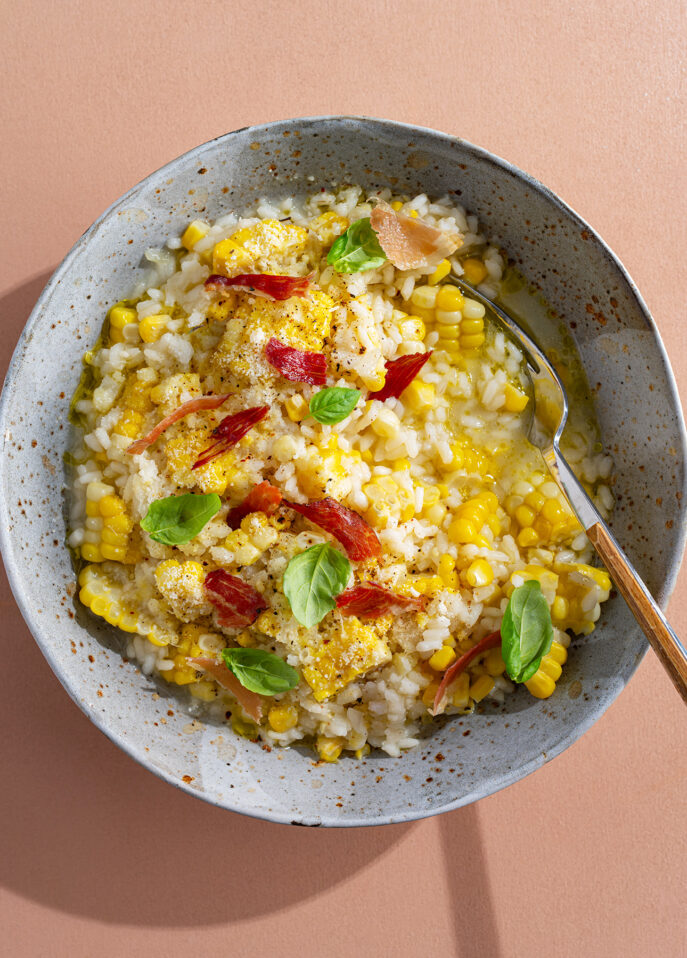 A bowl of creamy corn risotto topped with sliced red peppers and fresh basil leaves, with a spoon resting inside. The dish is served in a speckled ceramic bowl on a peach-colored surface. // FoodNouveau.com