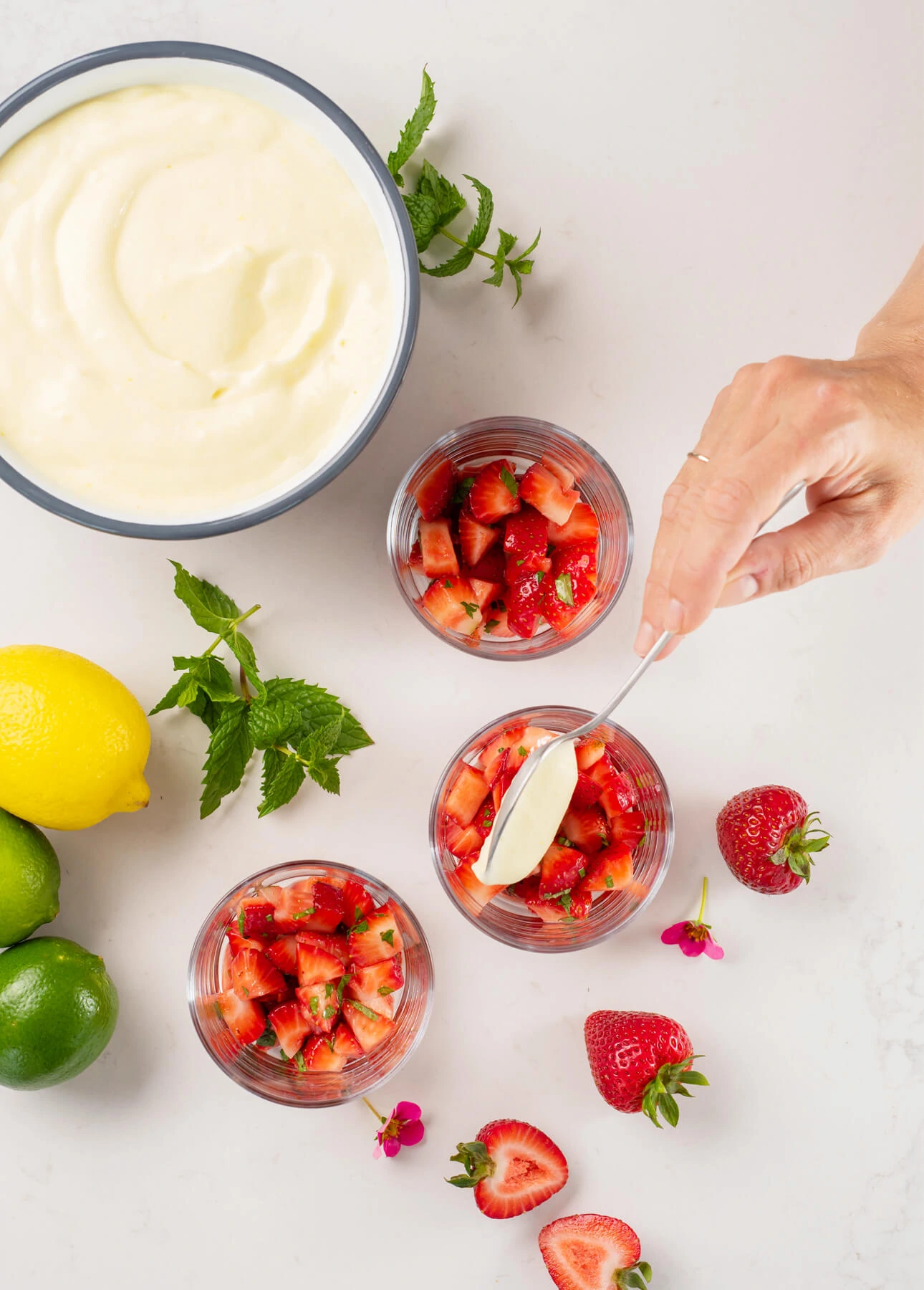 A hand spoons creamy citrus mousse over diced strawberries in glass cups, surrounded by whole strawberries, lemon, limes, mint leaves, and a bowl of cream on a white surface. // FoodNouveau.com