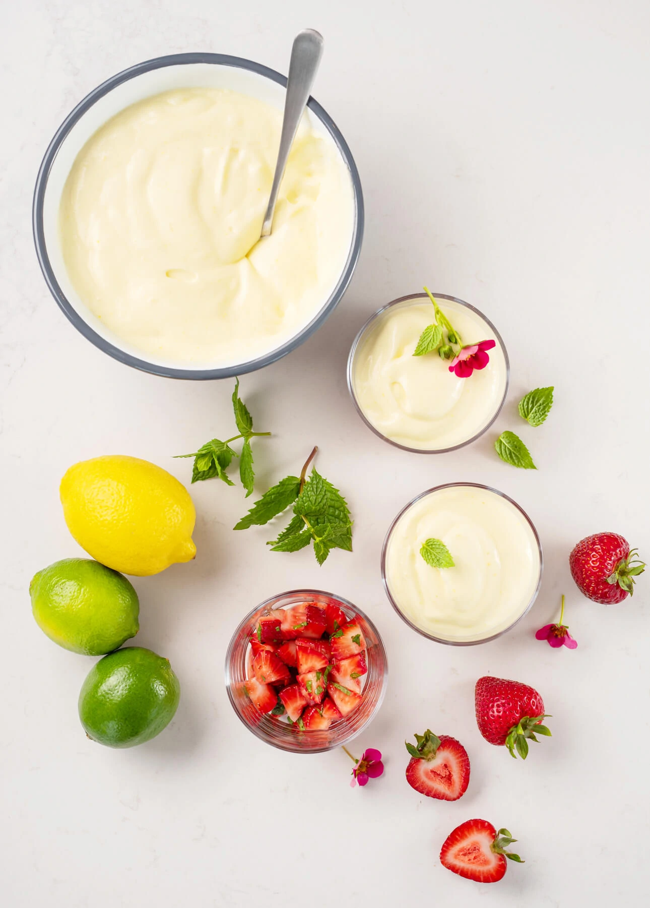 A bowl and small glasses of creamy citrus mousse are arranged with whole lemons, limes, mint leaves, strawberries, a dish of diced strawberries, and small pink flowers on a white surface. // FoodNouveau.com