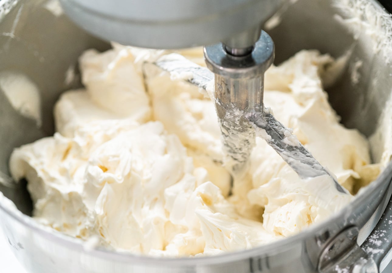 A close-up of thick, creamy frosting being mixed in a stand mixer with a paddle attachment. The frosting, perfect for vegan carrot cupcakes, is light in color and has a smooth, fluffy texture inside a metal mixing bowl. // FoodNouveau.com