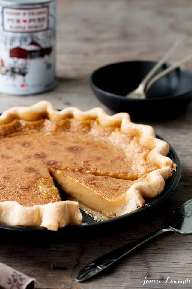 A creamy custard pie with a golden-brown top sits in a pie pan, missing one slice. The slice rests beside the pan, showing the smooth, light filling&mdash;a tempting choice for those who love classic maple dessert recipes. A tin and two forks are in the background. // FoodNouveau.com
