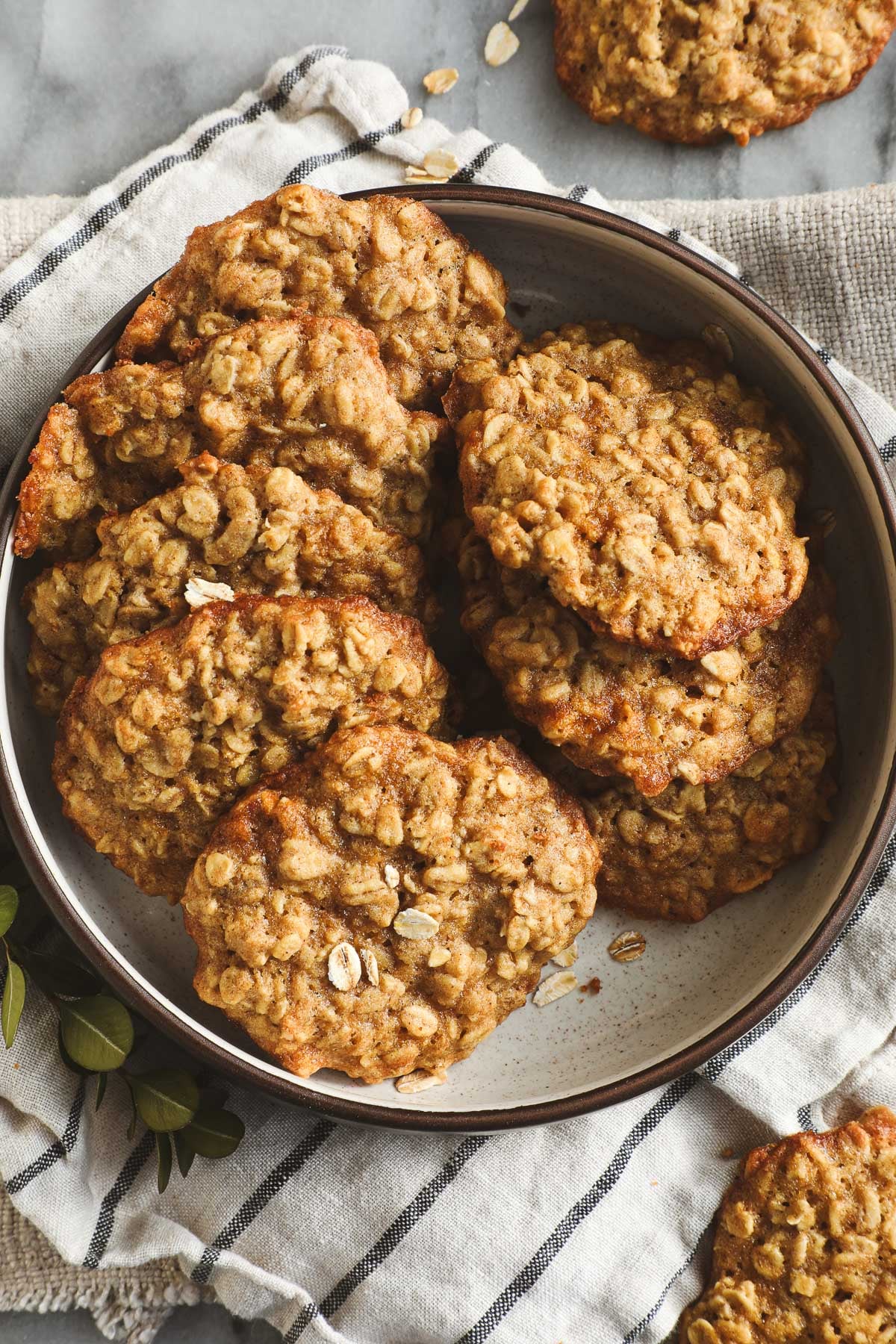 A plate filled with homemade oatmeal cookies sits on a striped cloth napkin; some cookies are stacked, showing their golden-brown, textured surface with visible oats&mdash;perfect for anyone seeking maple dessert recipes. // FoodNouveau.com