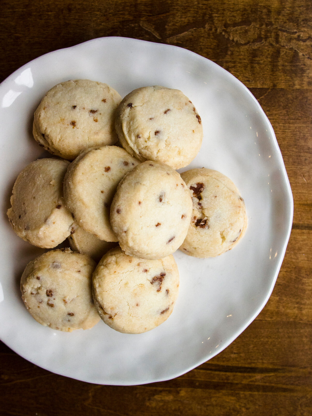 A white plate filled with round, light-colored cookies containing small bacon pieces, displayed on a wooden table&mdash;perfect for those seeking new maple dessert recipes. // FoodNouveau.com