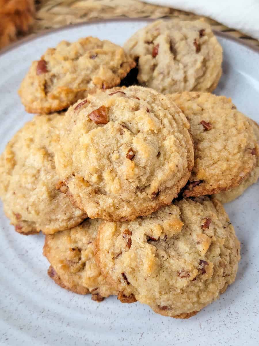 A plate of homemade maple pecan cookies with a golden-brown color and visible chopped nuts on top, stacked on a white ceramic plate&mdash;perfect for those seeking maple dessert recipes. // FoodNouveau.com