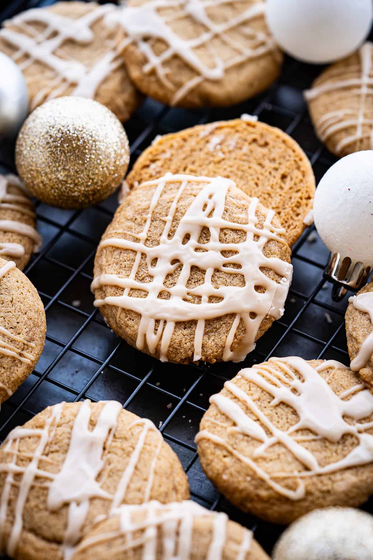 A close-up of round maple brown sugar cookies with white icing drizzle, arranged on a black cooling rack with gold and white Christmas ornaments nearby&mdash;a festive touch for those who love maple dessert recipes. // FoodNouveau.com