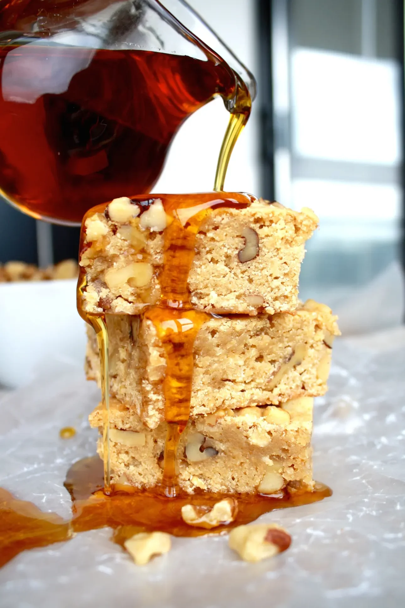 A close-up of syrup being poured over a stack of three nut-studded blondies on parchment paper, with extra syrup and chopped nuts around the base&mdash;perfect inspiration for maple dessert recipes. // FoodNouveau.com