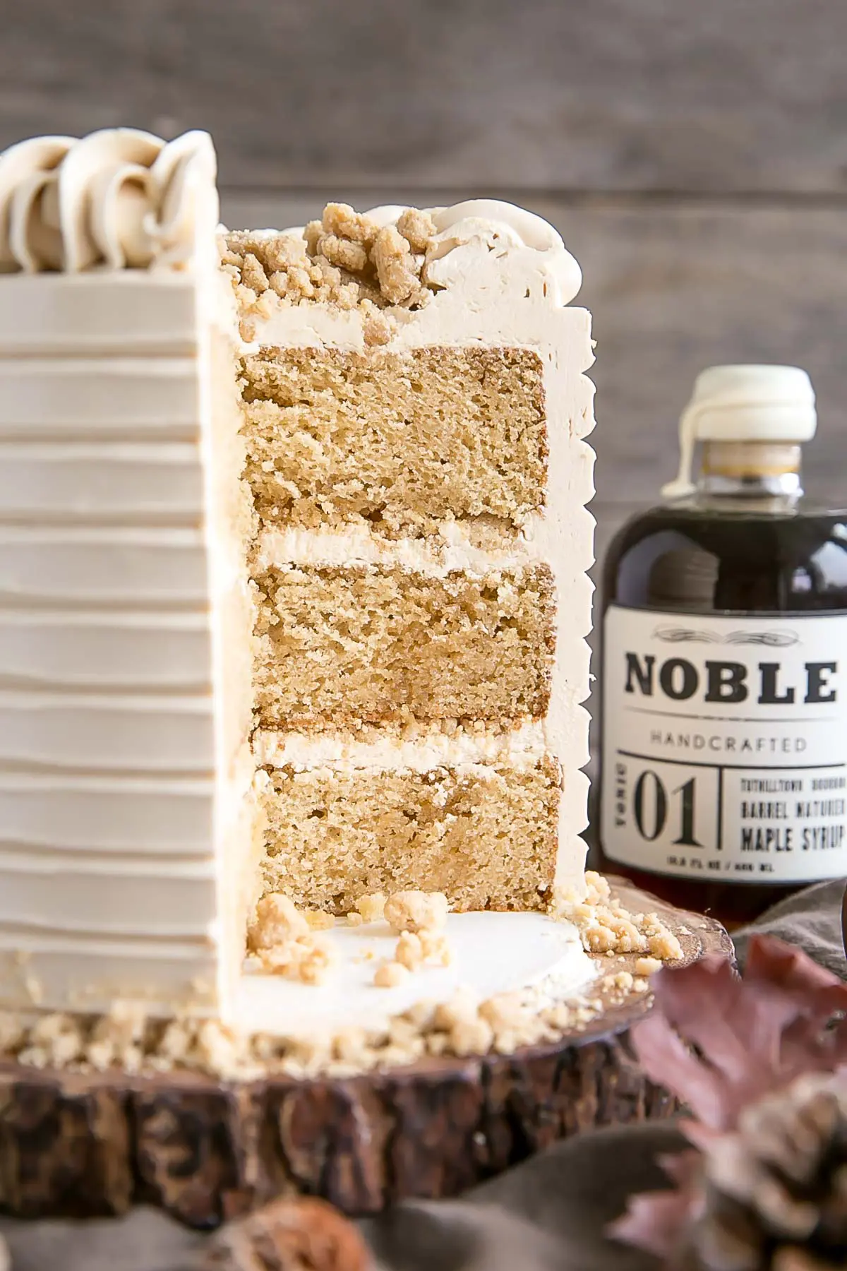 A three-layer cake with light brown frosting is sliced to reveal its interior. The cake sits on a wooden board with crumb topping&mdash;perfect inspiration for maple dessert recipes. A bottle labeled Noble Handcrafted Maple Syrup is in the background. // FoodNouveau.com