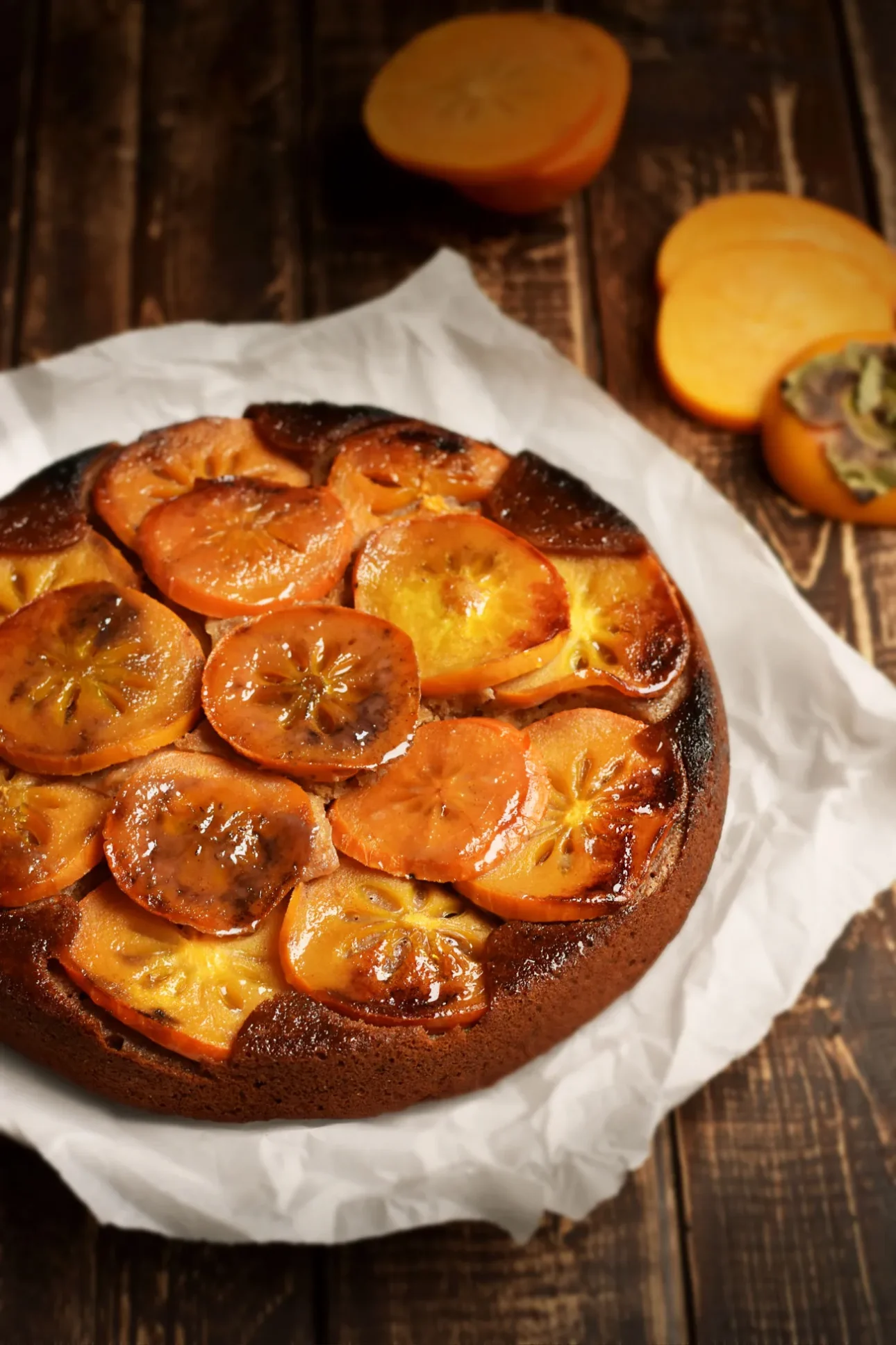 A round upside-down cake topped with caramelized persimmon slices sits on white parchment paper, perfect for fans of maple dessert recipes, with fresh persimmons and slices in the background on a rustic wooden surface. // FoodNouveau.com
