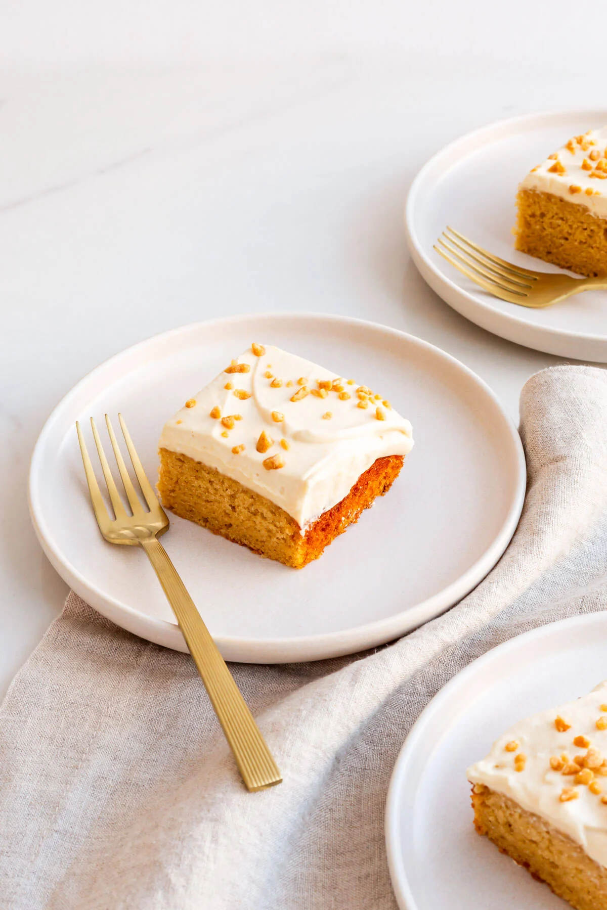 A square slice of maple cake with white maple frosting and nut sprinkles on a white plate, next to a gold fork. Two more similar slices are on separate plates in the background&mdash;perfect for those seeking maple dessert recipes. // FoodNouveau.com