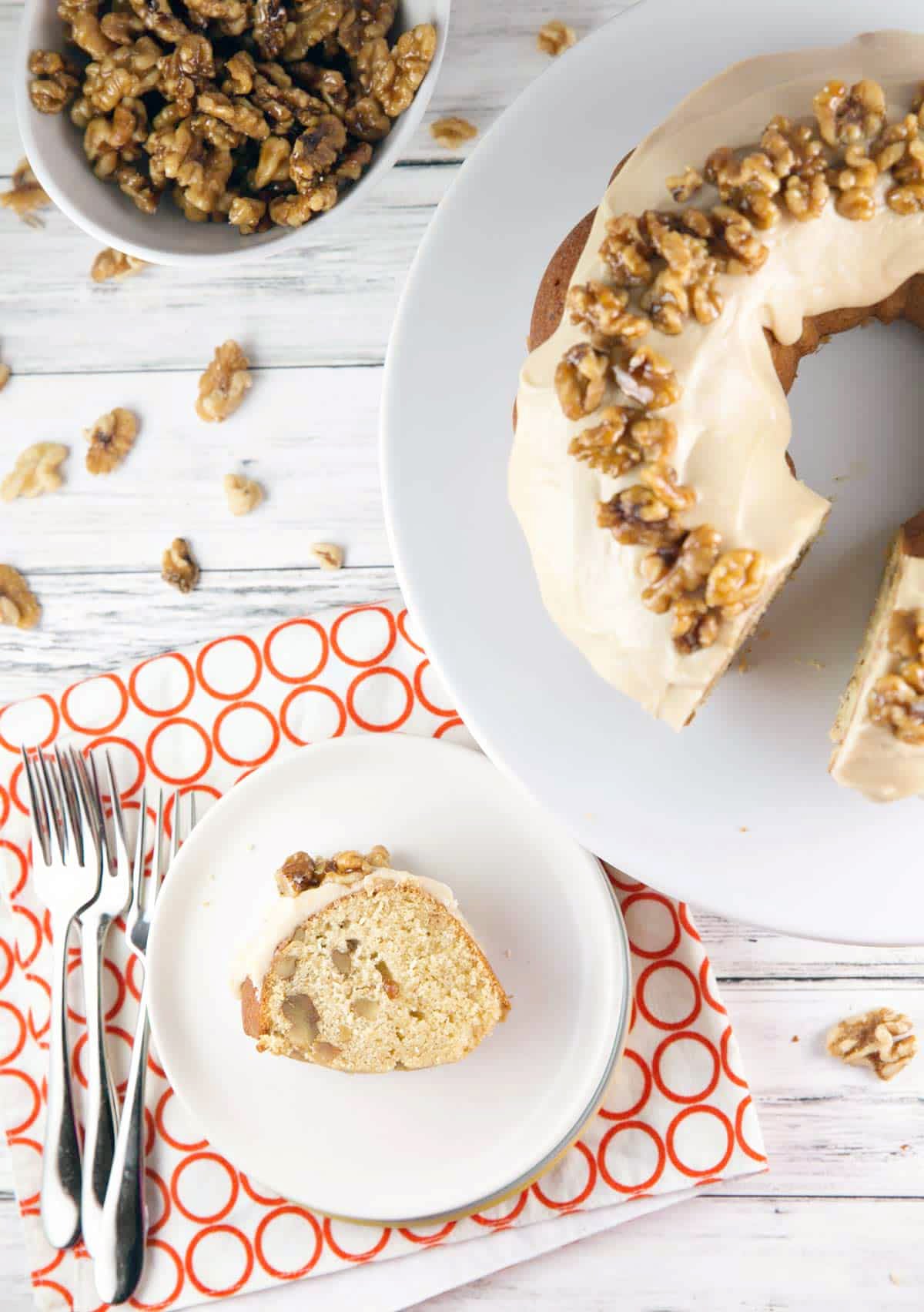 A sliced bundt cake with icing and walnuts sits on a white cake stand. A piece of this maple dessert recipe rests on a plate beside three forks, with a bowl of walnuts and scattered nuts on a white wooden table. // FoodNouveau.com