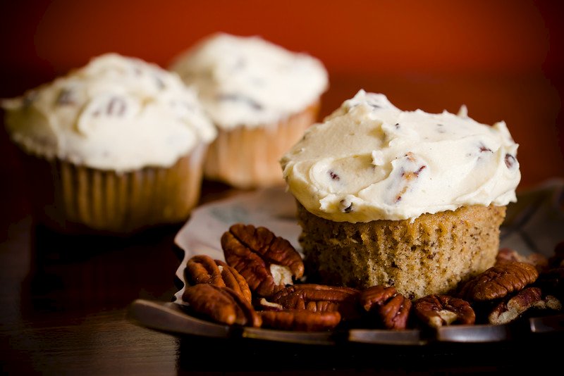 A close-up of a maple pecan cupcake with creamy frosting, surrounded by whole pecans on a plate. Two more frosted cupcakes, perfect for maple dessert recipes, are blurred in the background, all set against a dark surface and orange backdrop. // FoodNouveau.com