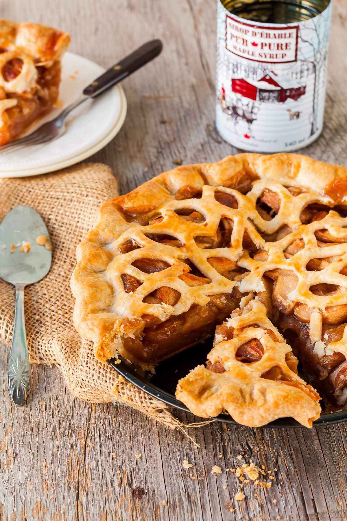 A lattice-topped maple apple pie with a slice removed sits on a rustic wooden table. The slice is on a plate nearby, and a can of maple syrup and a spoon are next to the pie&mdash;perfect inspiration for maple dessert recipes. // FoodNouveau.com