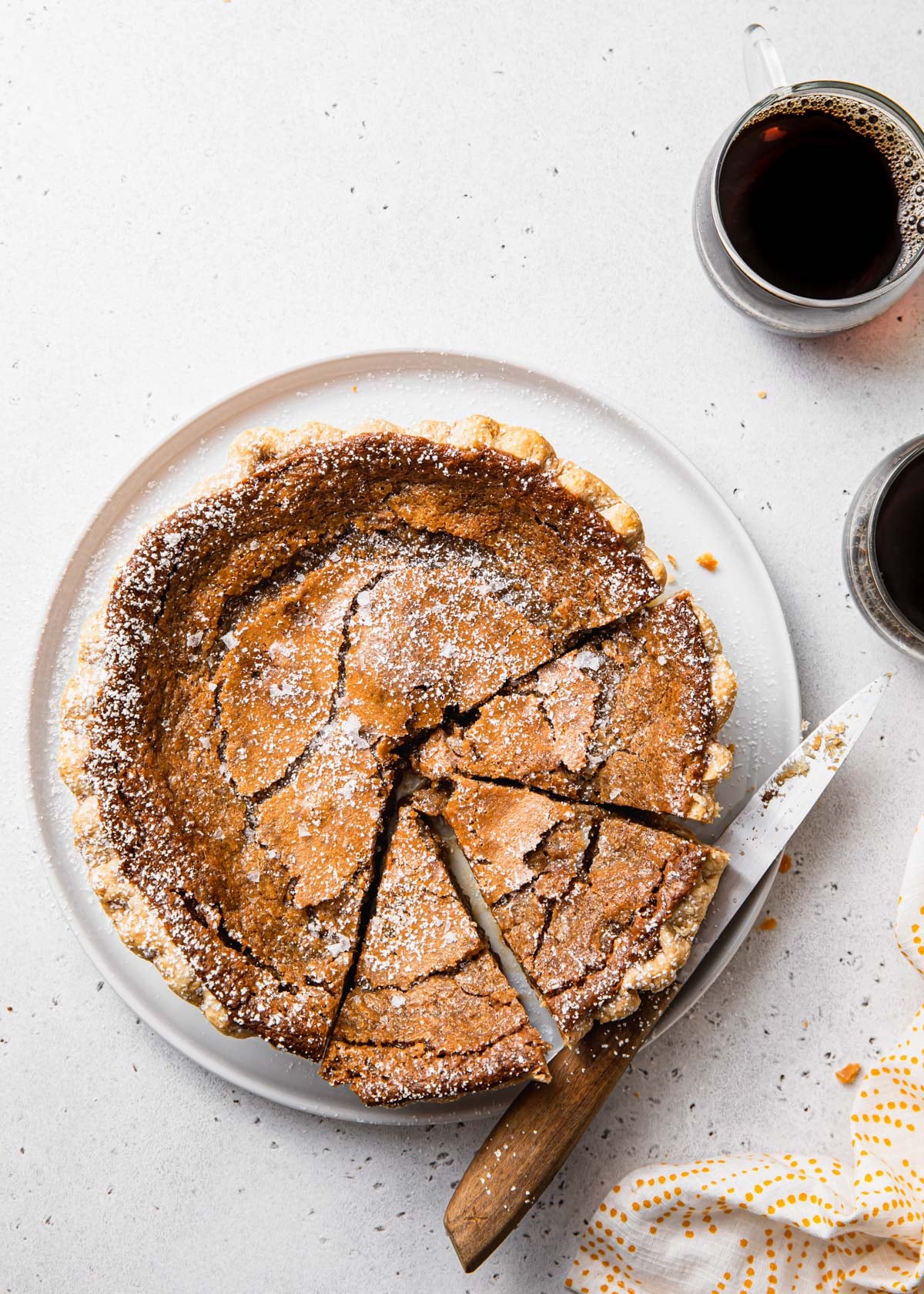 A partially sliced pie dusted with powdered sugar sits on a white plate with a knife, evoking cozy maple dessert recipes. Two mugs of black coffee are nearby on a light-colored surface with a white and yellow towel. // FoodNouveau.com