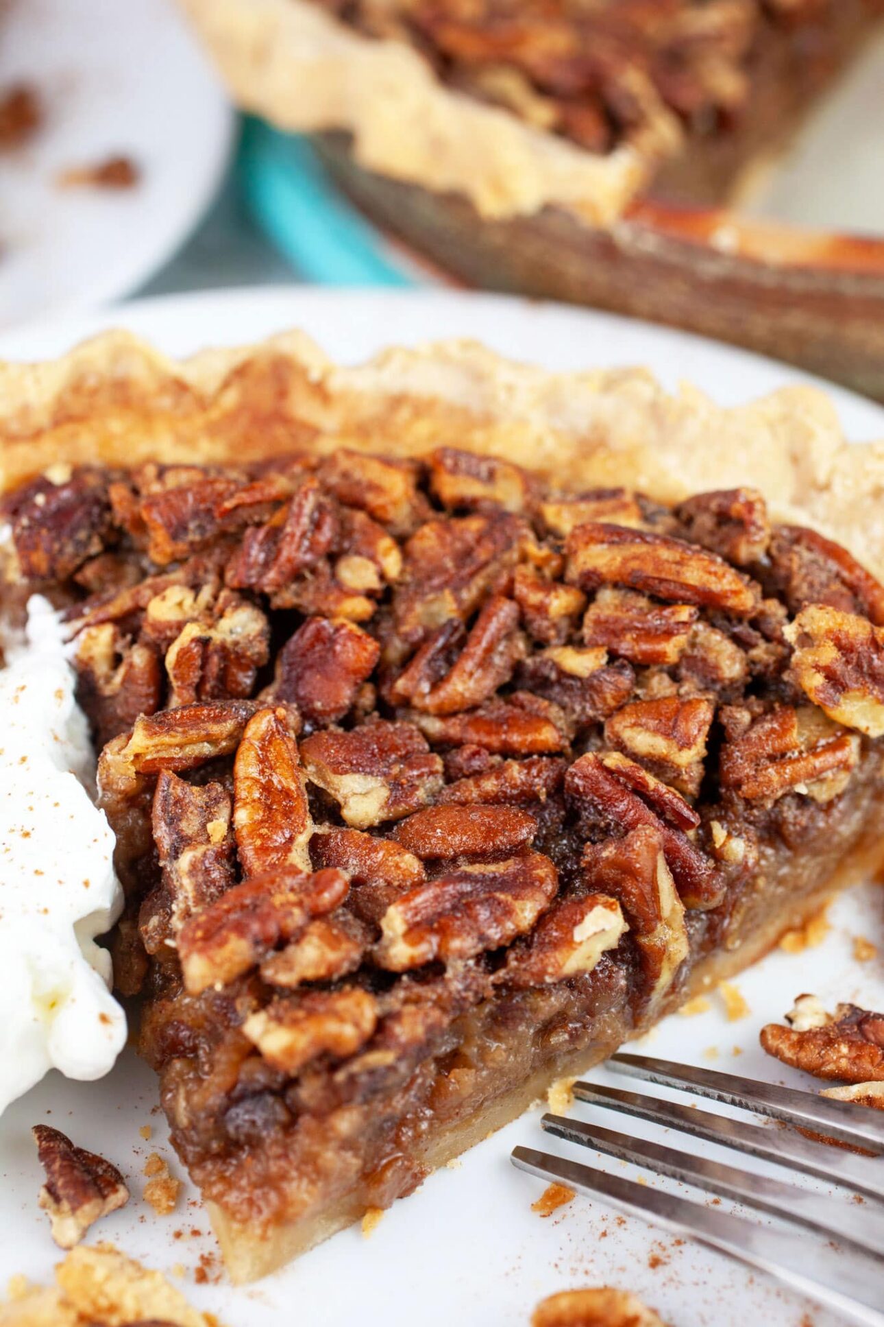 A close-up of a slice of maple pecan pie on a white plate, topped with crunchy pecans and served with a dollop of whipped cream on the side. Perfect for fans of maple dessert recipes, a fork rests nearby, ready to dive in. // FoodNouveau.com