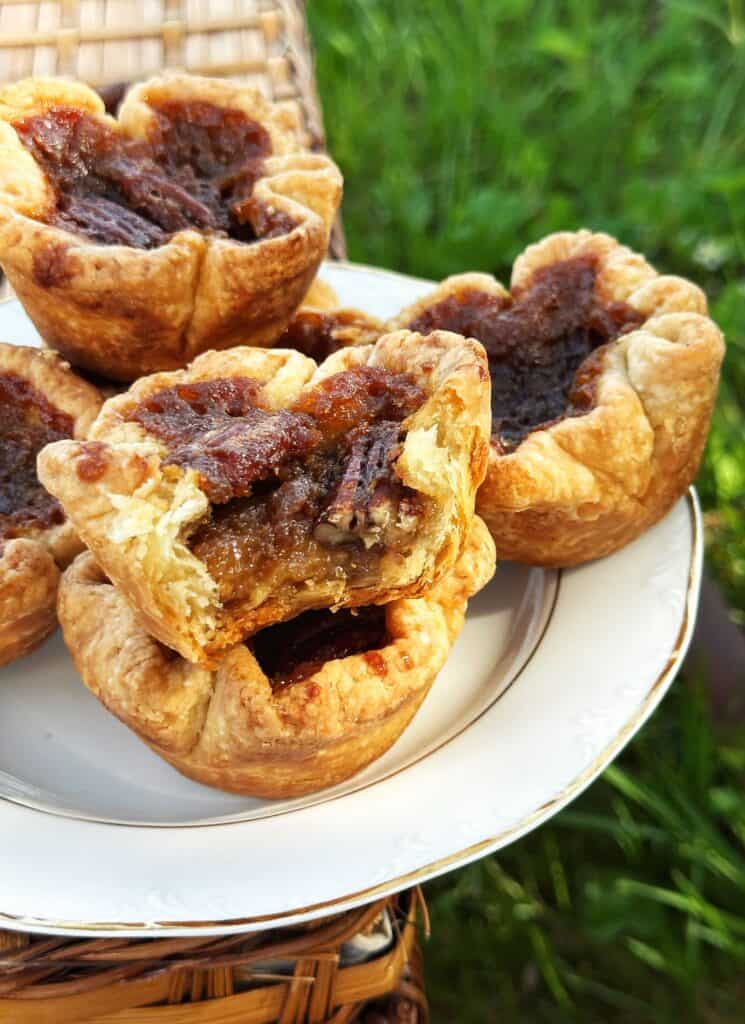 A plate of four maple pecan butter tarts, inspired by classic maple dessert recipes, sits outdoors on a wicker surface. Their flaky pastry and gooey, caramelized filling are visible, with one tart bitten into. Green grass is blurred in the background. // FoodNouveau.com