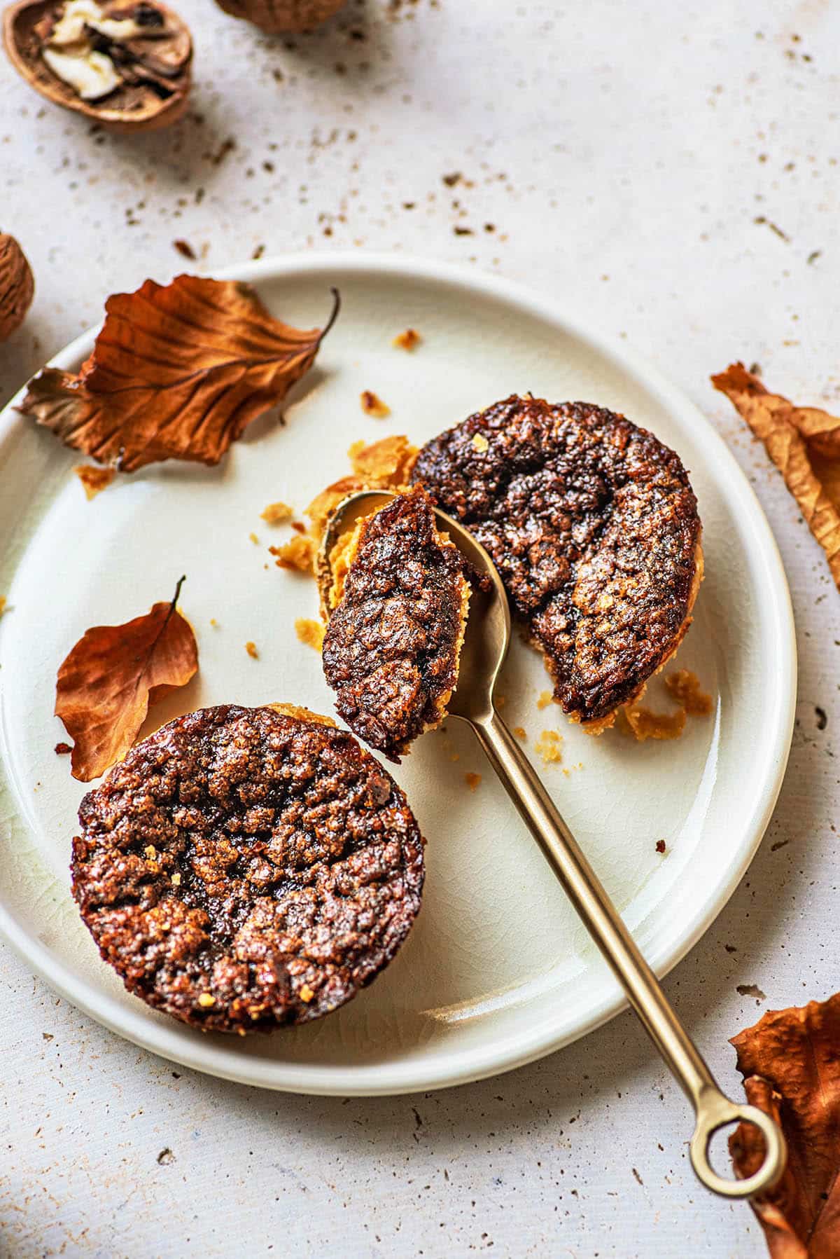 Two mini butter tarts on a white plate with a gold spoon cutting into one. Crumbs and dried autumn leaves are scattered around, evoking cozy vibes&mdash;perfect for those seeking new maple dessert recipes this fall. // FoodNouveau.com