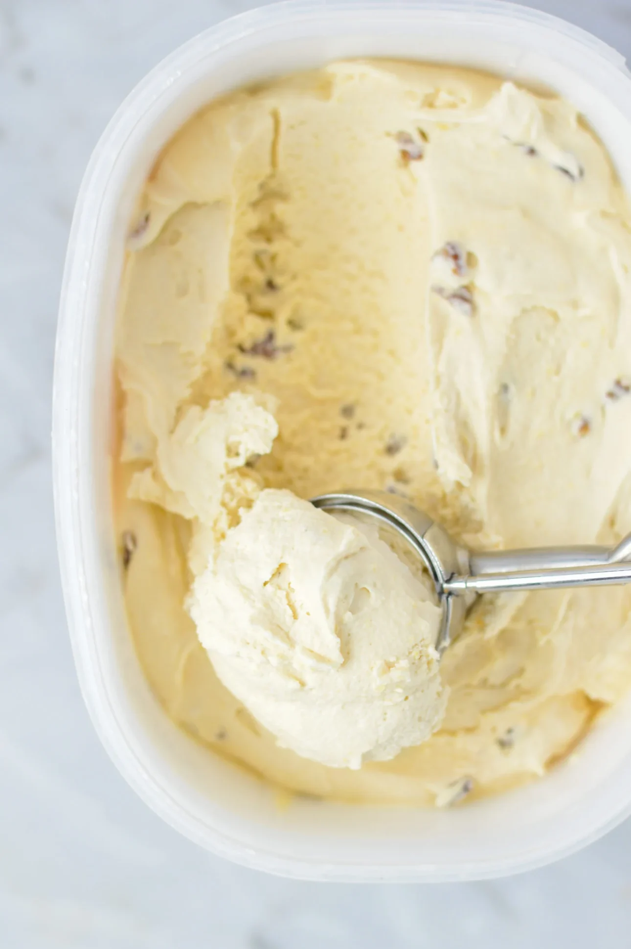 A metal ice cream scoop lifts a scoop of creamy maple ice cream with small mix-ins from a white plastic container on a light-colored surface, perfect inspiration for maple dessert recipes. // FoodNouveau.com